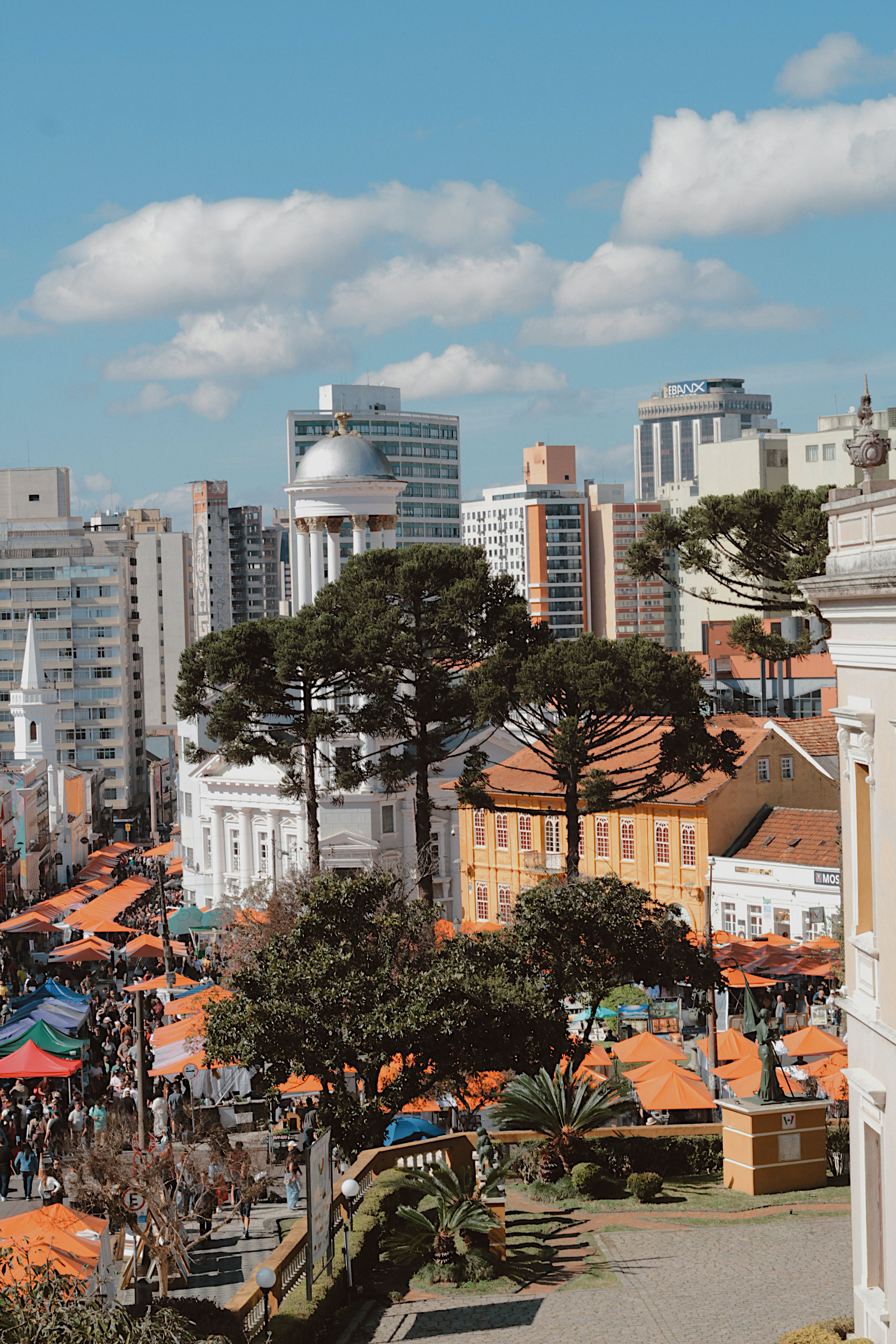 A vibrant aerial shot of Curitiba's city market with orange tents amid urban buildings and lush araucaria trees.
